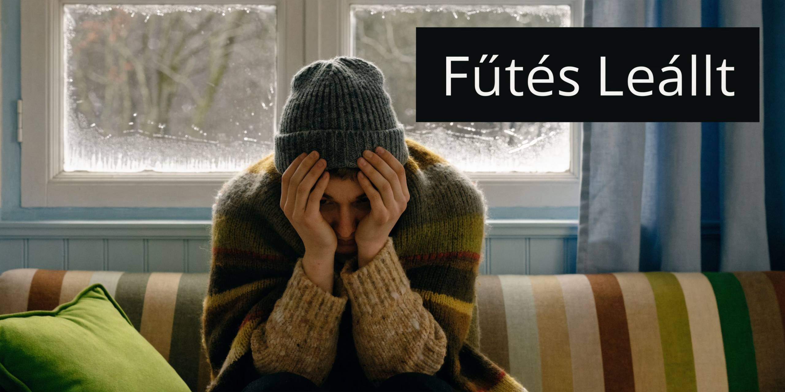 Egy fiatal férfi téli ruhában, takaróval betakarózva ül a hideg szobában, mert meghibásodott a fűtési rendszere.